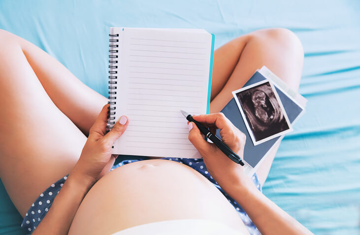 Pregnant woman makes notes in notebook and holding ultrasound image and medical documents at home interiors. Pregnancy, parenthood, preparation and expectation concept. Close-up, copy space, indoors.