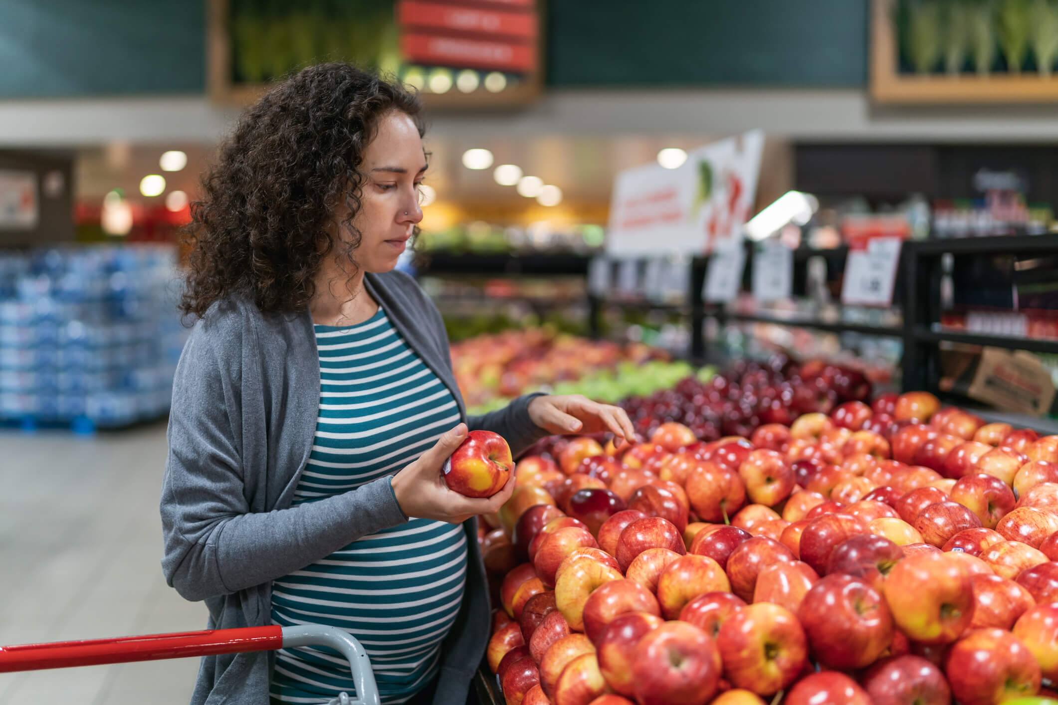 A young mixed race pregnant woman is in the produce section at the grocery store. The healthy woman is buying apples. She is wearing casual maternity clothing.