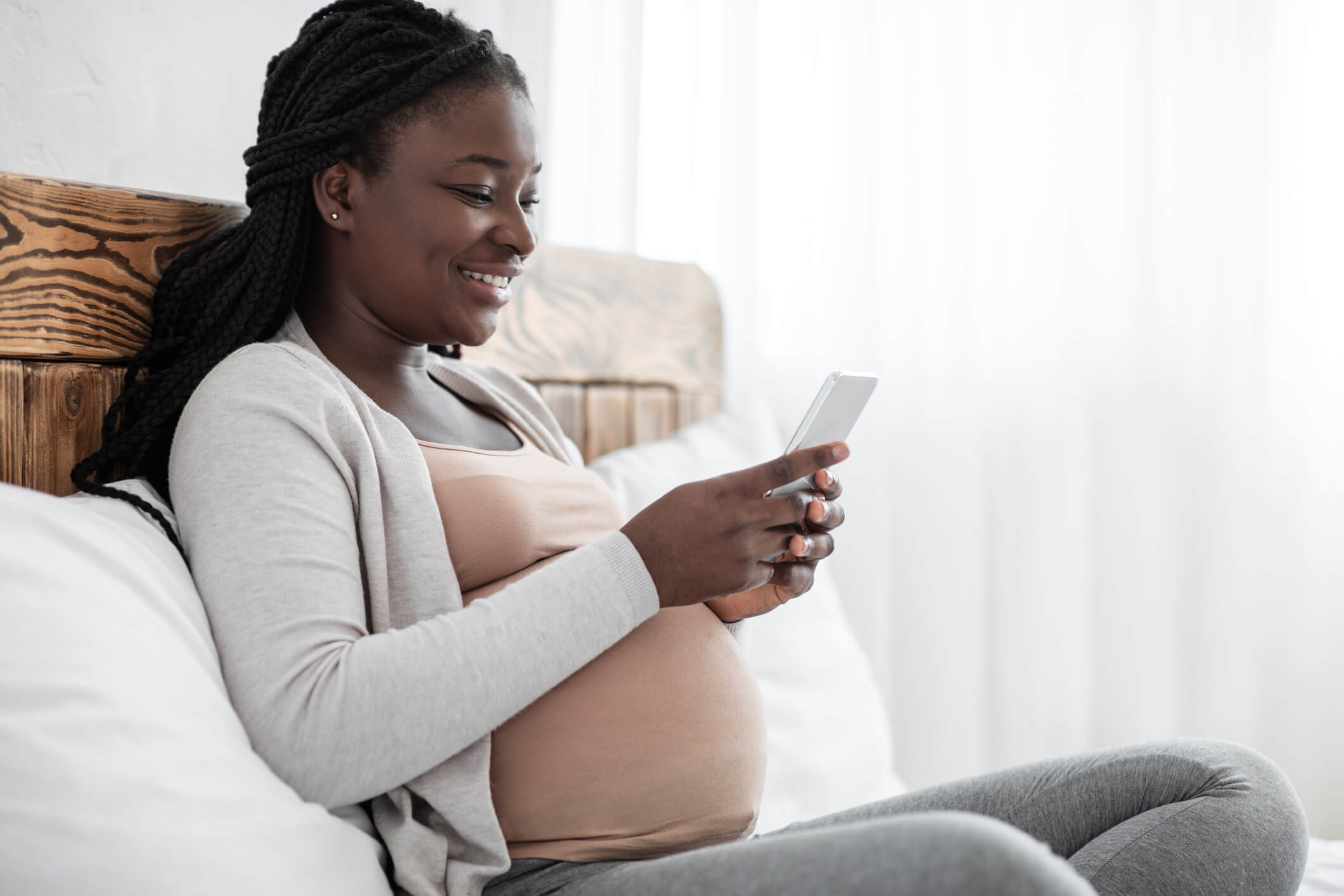 Joyful Black Pregnant Lady Spending Time With Smartphone At Home, Browsing Internet Or Messaging With Friends, African Expectant Woman Relaxing On Comfortable Bed, Side View Shot With Copy Space